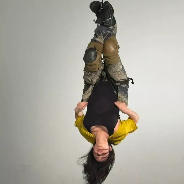 A picture shows Billie hanging from a rope while wearing camouflage pants, a black bodysuit, and yellow shrugs.