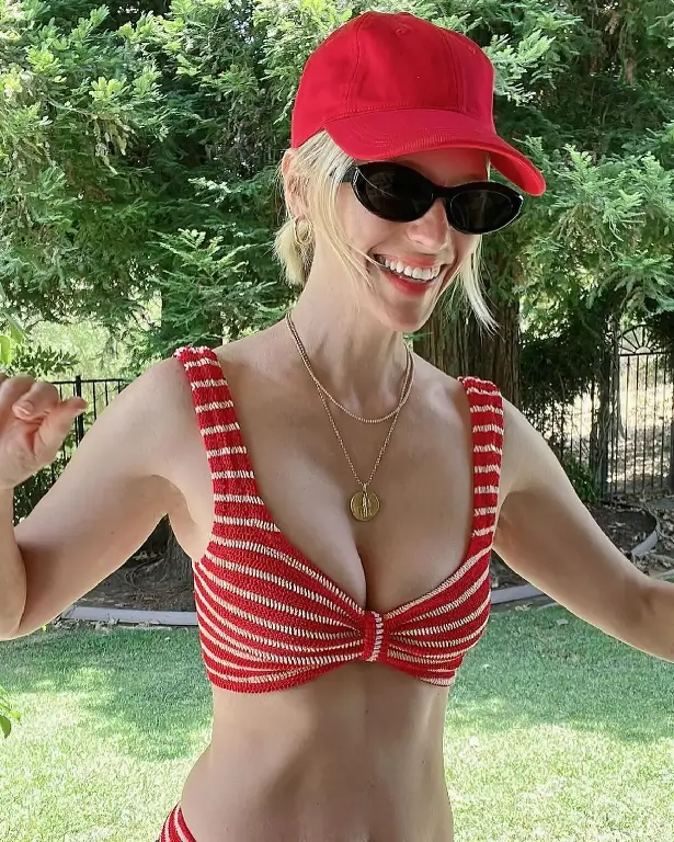 January in a tiny red and white bikini top tied at her front with high waisted bottoms, leaning forward in a wicker chair holding onto the side while beaming from ear to ear.