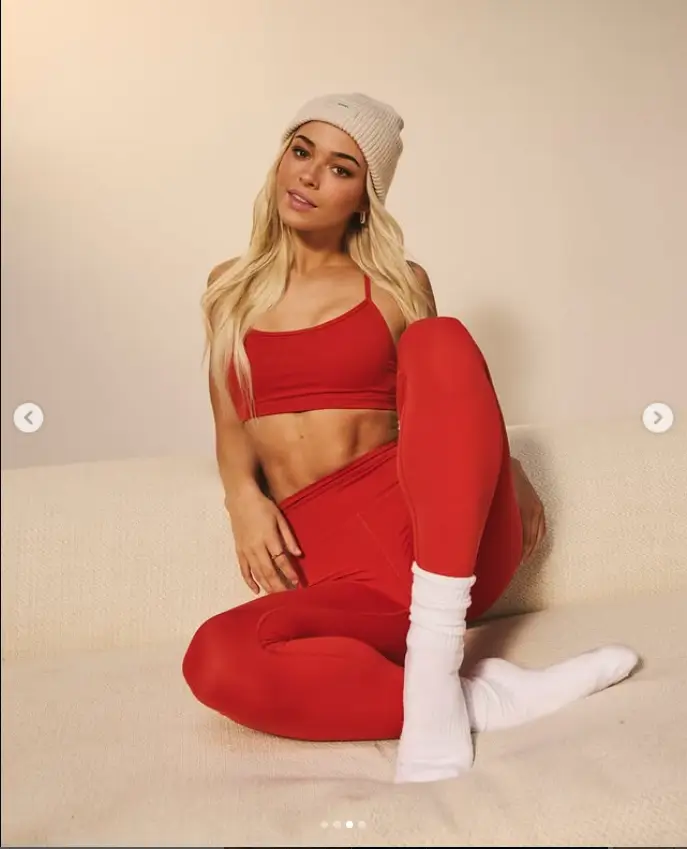 Her outfit features a cozy beige hat, a snug strap top, and eye-catching red leggings that beautifully complement her toned physique.